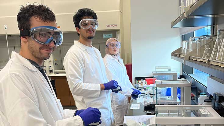 Three students in a chemistry lab in saftey gear and working with experimental equipment